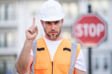 Güvenlik yeleği ve miğferli inşaat işçisi ellerini ileri uzatarak dur işaretinin yanında dur diyor. Erkek denetmen konut inşaatında güvenliği sağlıyor. Sıkı dikkat atmosferi, kentsel