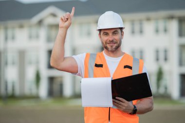 Güvenlik kasklı inşaat yöneticisi parmak kaldırıyor, klasörü ve evrakları açık tutuyor, proje detaylarını gözden geçiriyor. Erkek mühendis yerleşim yerinde. Odaklanmış yönetim atmosferi, modern konut