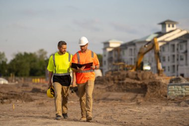 Koruyucu miğferler ve yansıtıcı yelekli iki erkek mühendis inşaat alanında el sıkışmaya hazırlanıyor. Sahne müzakere, karşılıklı saygı, profesyonel iletişim, proje ve takım çalışmasını gösteriyor.