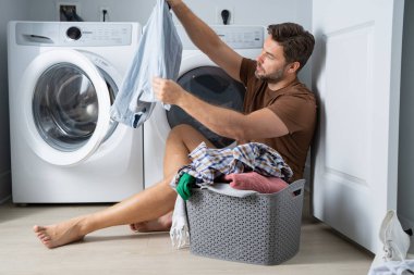 Çamaşır odasında çamaşır makinesinin yanında giysileri sıralayan mutlu bir adam. Adam kirli çamaşırlarıyla çamaşır makinesinin yanında bir evin zemininde oturuyor. Bir adam çamaşırlarını evde yıkıyor. Kirli çamaşırları olan bir adam.