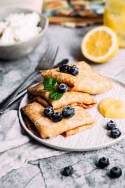 Ev yapımı süzme peynirli krep, yaban mersini ve gri tabakta limon ezmesi.