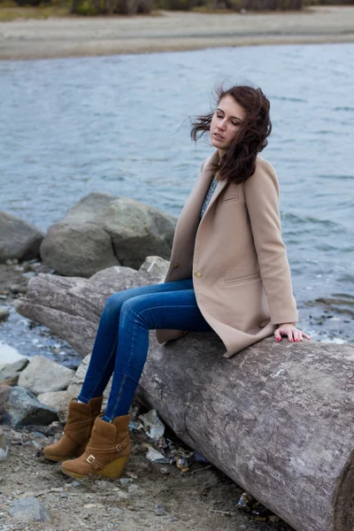 Stylish girl in autumn coat sitting by the water — Stock Photo, Image