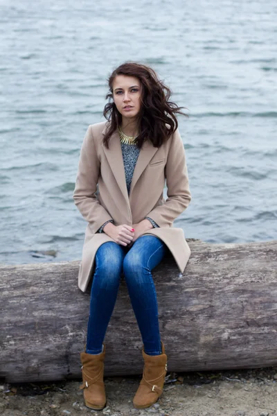 Stylish girl in autumn coat sitting by the water — Stock Photo, Image