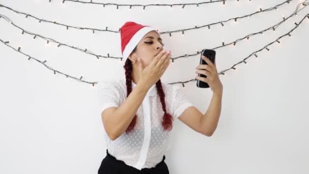 年轻姑娘戴着一顶红色圣诞帽 拿着手机打了一个视频电话 她高兴极了 一边跳舞 一边挥手 一边亲吻 — 图库视频影像