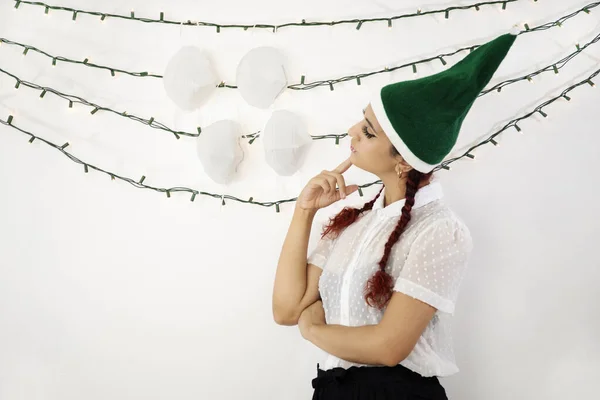 小女孩头戴绿色圣诞帽 头戴辫子 红头发 看着挂在一些花环上的面具 决定着该戴哪一顶 — 图库照片