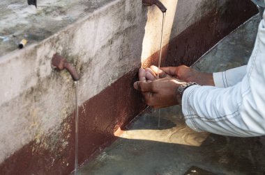 Musluktan kirli bir gölete düşen su, suyu aydınlatan bir ışık hüzmesi, suda ellerini yıkayan insan, sadece eller görünür, güneşli bir gün.