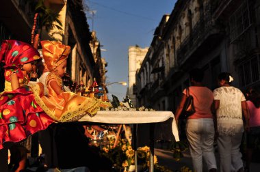 İki dindar bebek, biri sarı, diğeri kırmızı giyinmiş, dini şeyler ve çiçeklerle dolu bir masada, kadınlar eski bir şehrin sokaklarında yürüyorlar, Afro-Küba dini, güneşli bir gün.