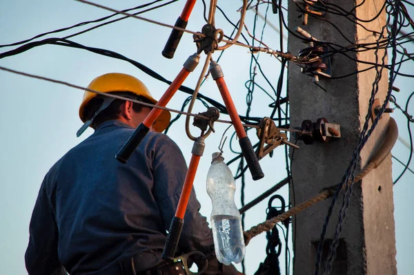 Sarı bir miğfer takmış elektrik hattı çalışanı, sokak kablolarında kelepçelerle çalışıyor. Kemerlerle korunan bir direğin üstünde.