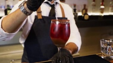 Barmen tezgahta kokteyl hazırlıyor. Kristal bardakta metal karıştırıcı şarap ve demirhindi suyuyla karıştırılıyor. Ellerine siyah eldivenlerle yakın plan çek..