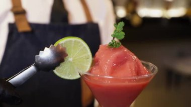 Barmen tezgahta kokteyl hazırlıyor. Metal bir klemple cam bir kadehe limon dilimi ve daiquiri kokteyli koymak. Ellerine siyah eldivenlerle yakın plan çek..