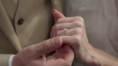 Yakın plan beyaz çift alyanslarını değiş tokuş ediyor, damat bandından gelin parmağına kayıyor, eller duygudan titriyor, bej takım elbise ve peçe.