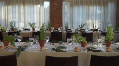 Kapalı masa ziyafeti, masa süslemeleri, tuğla duvar ve uzun pencereler, sıra sıra sandalyeler, beyaz çarşaflar, yeşillikler ve yeşillikler.