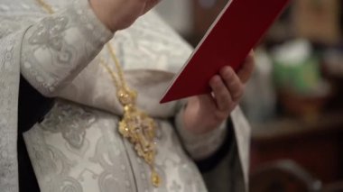 Kırmızı kitaptan edebiyat metni okuyan rahip süslenmiş altın haç ve desenli bornoz giyerken elleri ve cübbeyi yakından görüyor.