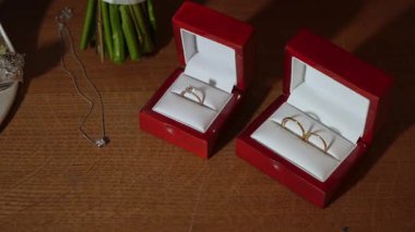 Masadaki kırmızı kutularda evlilik yüzükleri, Solitaire nişan yüzüğünün yakın aranjmanı ve buket buketlerinin yanında çift alyanslar.