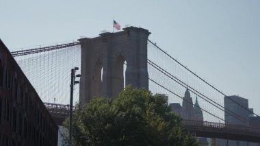 Brooklyn Köprüsü Kulesi Dalgalı Bayrak ve Ağaçlı Nehir Kıyısı Yumuşak Sabah Pusulası Taşı Kemerleri ve Askıya Alma Kabloları Sakin Tarih Oluştur