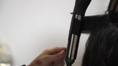 Saç stilisti, uzun kıvırcık, saç maşalı, elleri kıvırcık, elleri kapalı, salon aletleri görünür, odaklanmış teknikleri var.