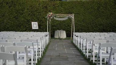 Beyaz sandalyeler çiçek kemerinin altındaki yeşil çimenlikte, banliyö çitlerinde ve arka planda, katlanan sandalyeler koridora dizilmiştir.,