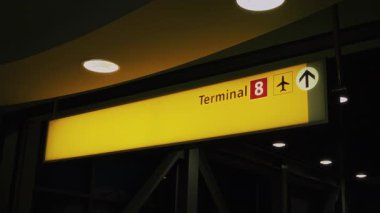Havaalanındaki Terminal 8 'i gösteren parlak ışıklı tabela, bir uçak simgesini ve yön okunu gösteriyor, karanlık bir iç arkaplan ve tepe ışıkları ile