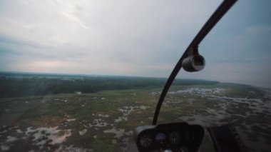 Helikopterin kokpiti yeşil sulak alanlara ve ağaçlara alçaldığını gösteriyor. Bulutlu bir gökyüzü uçuş sırasında dramatik bir zemin oluşturuyor.