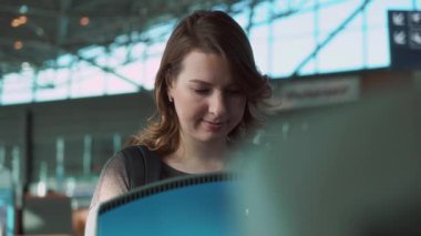 Açık kahverengi saçlı genç bir kadın, büyük pencerelerden gelen doğal ışıkla aydınlatılmış bir havaalanı terminalinde mavi bir bilet gişesiyle nişanlanıyor.