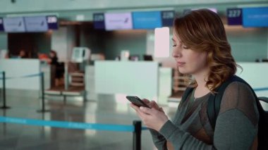 Beyaz kadın gezgin, çalışanlar ve dijital uçuş bilgileriyle çevrili bir havaalanı kontrol gişesinde akıllı telefonuyla etkileşime geçiyor.