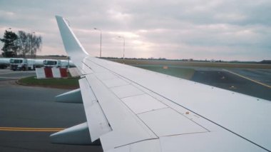 Uçak kanadı pistte taksiyle giderken pencereden görülebiliyor. Bulutlu bir gökyüzünün altında kanat detaylarını ve havaalanı çevresini vurguluyor.
