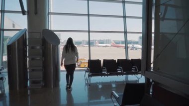 Beyaz gömlekli kadın havaalanı pencerelerine yaklaşır, pistteki uçaklara hayran kalır, modern koltuklar ve parlak güneş ışığı terminali aydınlatır.