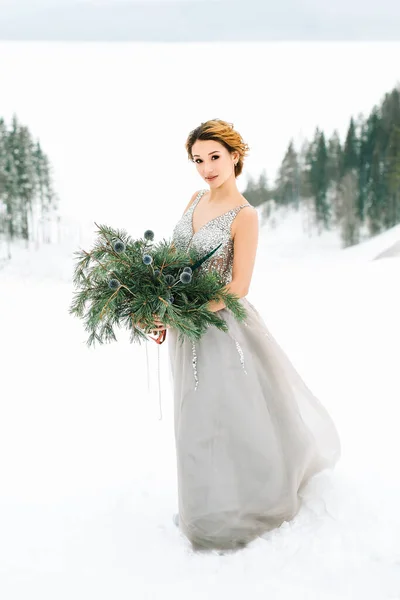 Wedding dress in the winter season. A bride with a haircut stands in a sparkling silver wedding dress on a snowy slope with a bouquet of pine trees. Christmas wedding concept. Selected focus. Blurred background