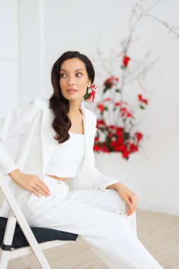 Şık ve zarif bir takım elbiseyle sandalyede oturan beyaz tenli esmer kadın portresi. Akşam ekipmanı, kutlama için kıyafet. Pantolonlu bir gelinlik, modern tarzda..
