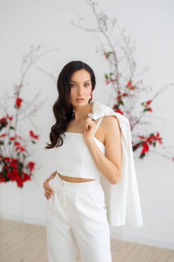 Şık ve zarif takım elbiseli, elinde ceket tutan beyaz, esmer bir kadın. Gelin, alışılmadık bir çiçek karışımının arka planında poz veriyor.