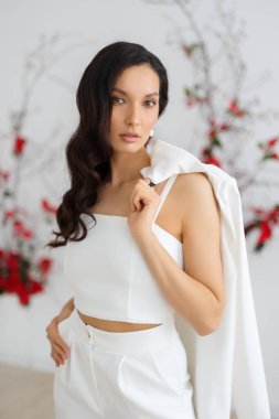 Şık ve zarif takım elbiseli, elinde ceket tutan beyaz, esmer bir kadın. Gelin, alışılmadık bir çiçek karışımının arka planında poz veriyor.