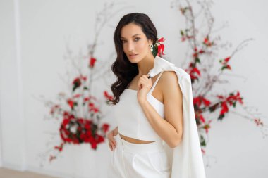 Şık ve zarif takım elbiseli, elinde ceket tutan beyaz, esmer bir kadın. Gelin, alışılmadık bir çiçek karışımının arka planında poz veriyor.