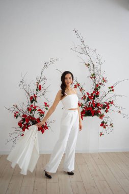 Beyaz pantolonlu gelin, özel bir düğün töreni için çiçek desenli alışılmadık bir kompozisyonun arka planında poz veriyor.