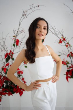 Beyaz pantolonlu gelin, özel bir düğün töreni için çiçek desenli alışılmadık bir kompozisyonun arka planında poz veriyor.