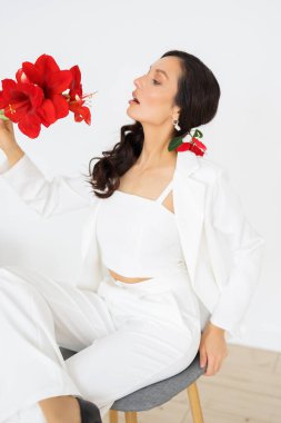 Beyaz pantolonlu, şık bir gelin ve bir buket kırmızı amarilllis sandalyede oturuyor. Düğün kış mevsiminde.