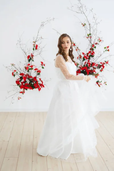 Bride in a white wedding dress poses against the background of an unusual composition of natural poinsettia flowers for a chamber wedding ceremony. Spins around in a dress and holds it in her hands.