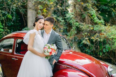 Eski arabanın ve tropikal bahçenin önünde damatla güzel bir gelin. Buket ellerde ve detaylarda. Kız mutlu, gülümsüyor ve dans ediyor. Modern tarzda bir gelinlik hayal et.