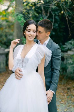 Zarif bir gelinlik içinde çekici bir gelin, sıcak bir yaz gününde, tropik bir bahçede gri bir takım elbise içindeki damadı öpüyor..