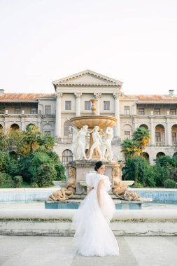 Parktaki fıskiyede poz veren muhteşem bir gelin, harika bir dantel elbise giyiyor. Gelinlik giymiş gülümseyen genç kadın kamerada poz veriyor.