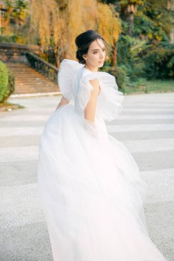 Gelinlik giymiş güzel ve havalı gelin rüzgârda kumaşla oynuyor. Güzel bir eş çeşmenin yanındaki parkta koşar. Moda ve düğün konsepti.