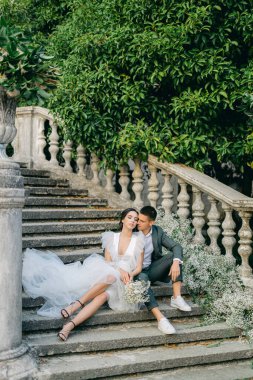 Gelin ve damat oturur ve çingene çiçekleriyle süslenmiş bir parkta taş bir merdivenin arka planında poz verirler. Düğün dekoru, beyazlı düğün konsepti..