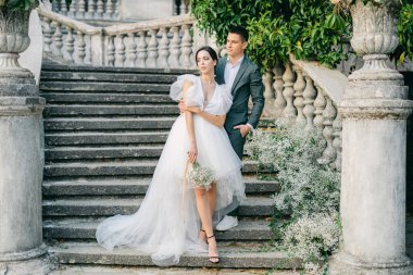 Şık takım elbiseli yakışıklı bir adamın beyaz gelinlik içindeki büyüleyici genç gelinini kucaklayan uzun bir portresi. Parkta beyaz çiçeklerle süslenmiş taş bir merdivenin arkasında poz veren mutlu yeni evli çift..