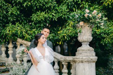 Şık takım elbiseli yakışıklı bir adamın beyaz gelinlik içindeki büyüleyici genç gelinini kucaklayan uzun bir portresi. Parkta beyaz çiçeklerle süslenmiş taş bir merdivenin arkasında poz veren mutlu yeni evli çift..