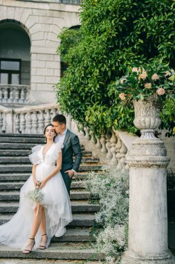 Şık takım elbiseli yakışıklı bir adamın beyaz gelinlik içindeki büyüleyici genç gelinini kucaklayan uzun bir portresi. Parkta beyaz çiçeklerle süslenmiş taş bir merdivenin arkasında poz veren mutlu yeni evli çift..