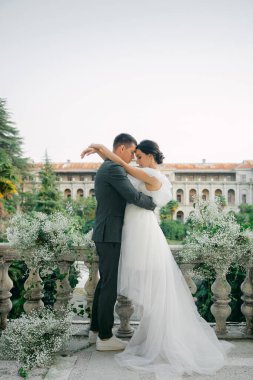 Muhteşem bir bahçe manzarasının arka planında şık bir çift kucaklaşması. Modaya uygun bir gelinlik içinde genç ve şık bir gelin ve yakışıklı bir damat düğün kemerinin önünde poz veriyor..