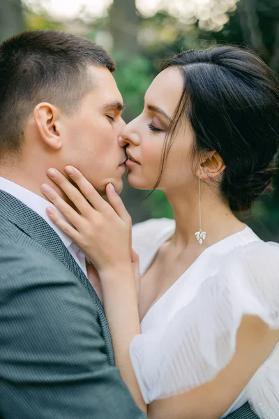 attractive bride in a delicate fashionable wedding dress kisses the groom in a gray suit near stone columns in a tropical garden on a warm summer day.