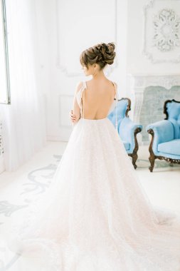 Güzel kız, gelin gelinlik içinde arkası açık duruyor. Yatak odası ve gelinin sabahı. Püsküllü giyin. Mavi klasik sandalyeli beyaz bir iç mekanda.