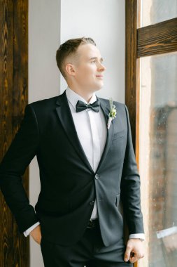 Siyah bir smokin ve papyon giymiş damat pencerenin arkasına poz veriyor. Modaya uygun klasik gelinlik ve koca imajı konsepti.