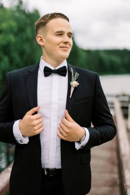 Orman gölünün ortasında bir rıhtımda duran smokinli mutlu genç bir kocanın portresi. Klasik Avrupa açık hava düğünü kavramı