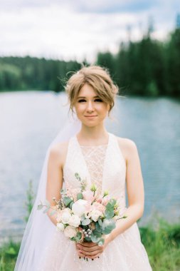 Orman Gölü 'nün önünde çiçek ve dekorla güzel bir gelin. Buket ellerde ve detaylarda. Kız mutlu, gülümsüyor ve dans ediyor. Modern tarzda bir gelinlik hayal et.